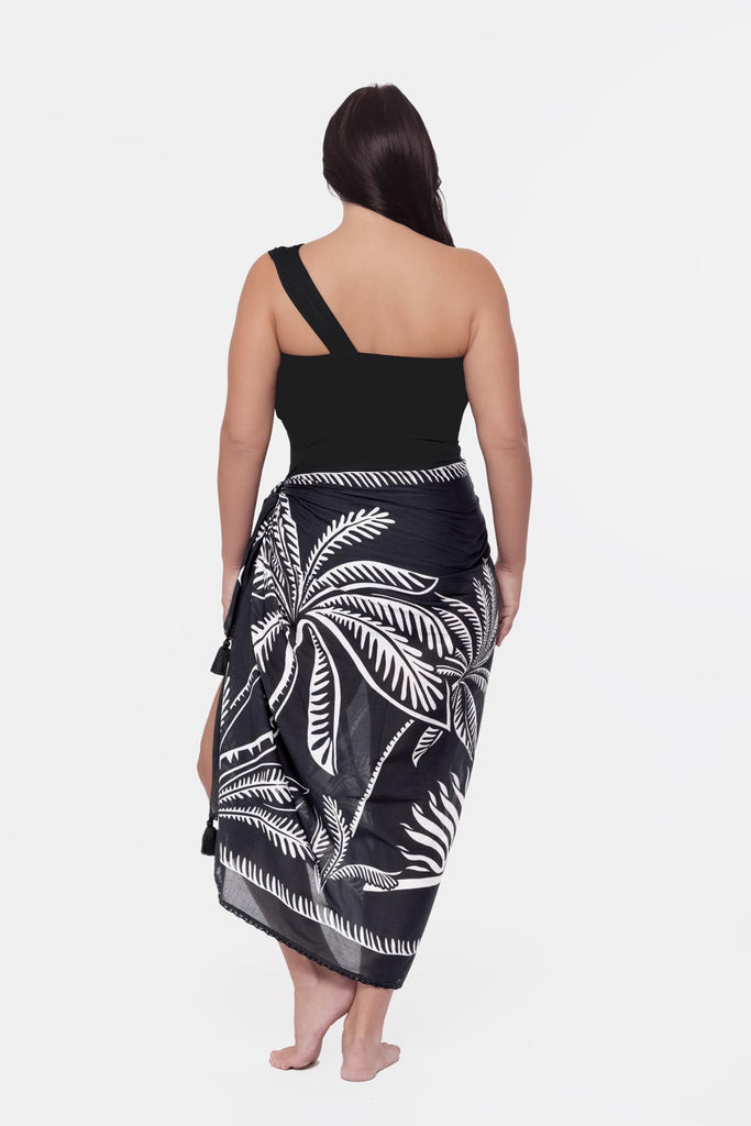 Woman turned away wearing a black one shoulder swimsuit with a palm leaf pattern cover up around the waist