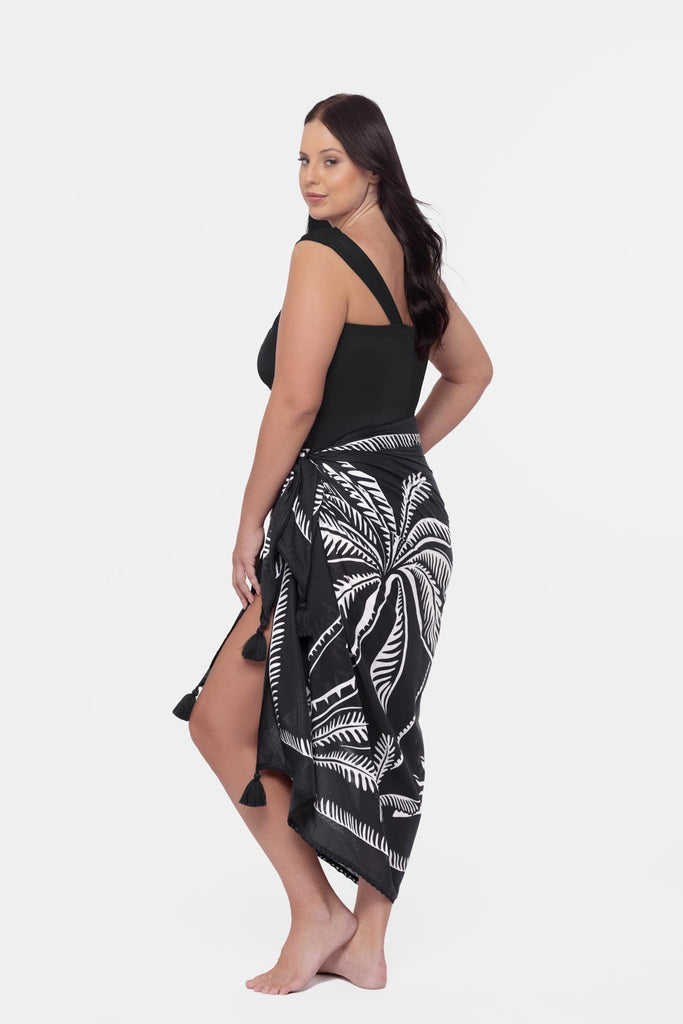 Woman turned left wearing a black one shoulder swimsuit with a palm leaf pattern cover up around the waist